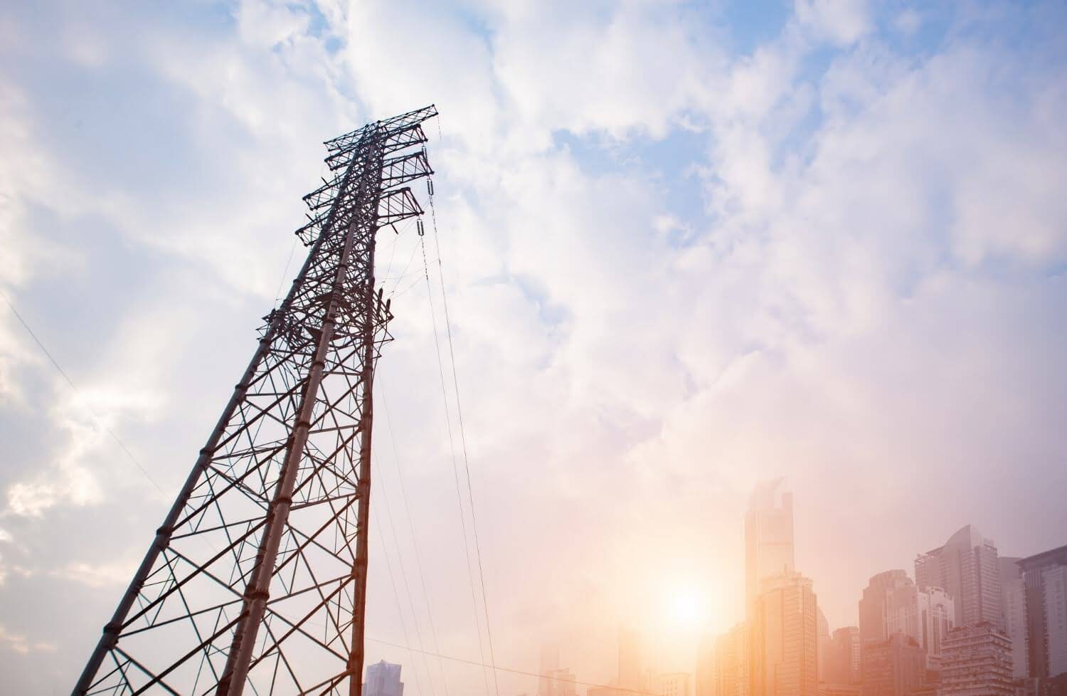 Torre de transmissão de energia elétrica vista de baixo, com o céu e cidade ao fundo.
