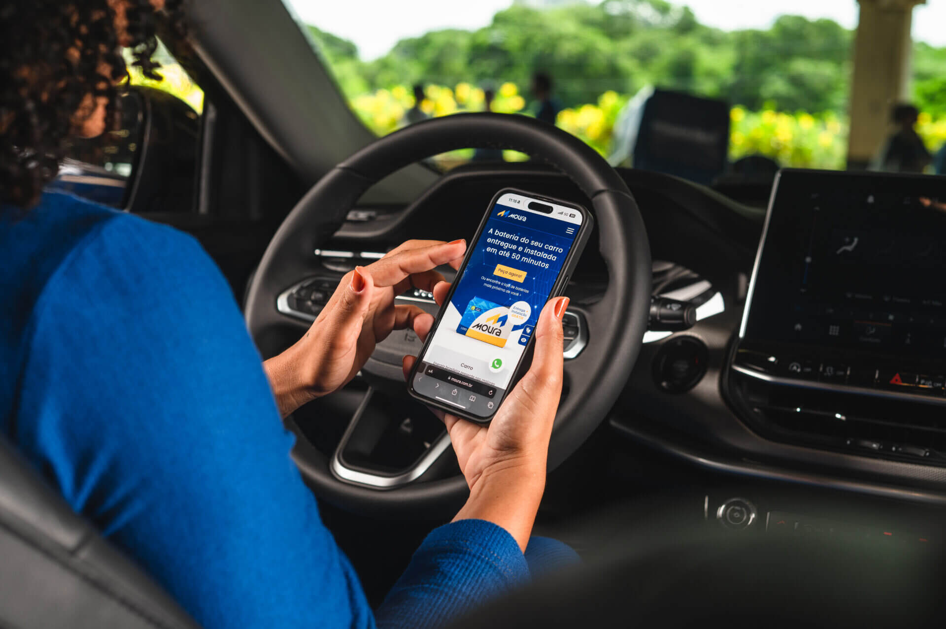 Mulher sentada no banco do motorista de um veículo, enquanto segura um smartphone com a tela do site do Moura Fácil.