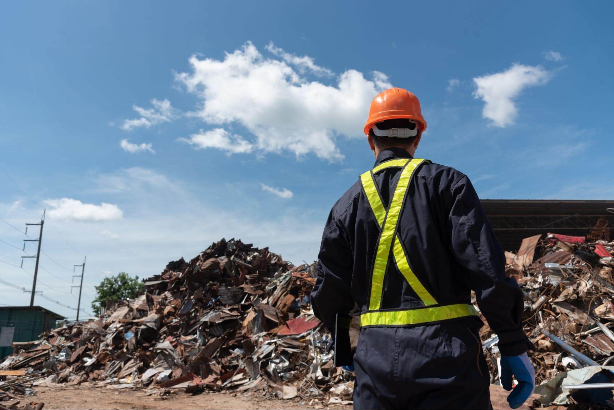 um homem que trabalha com o descarte de lixo veste um uniforme e capacete e permanece em pé em frente a uma junção de lixos, no que parece ser um aterro a céu aberto