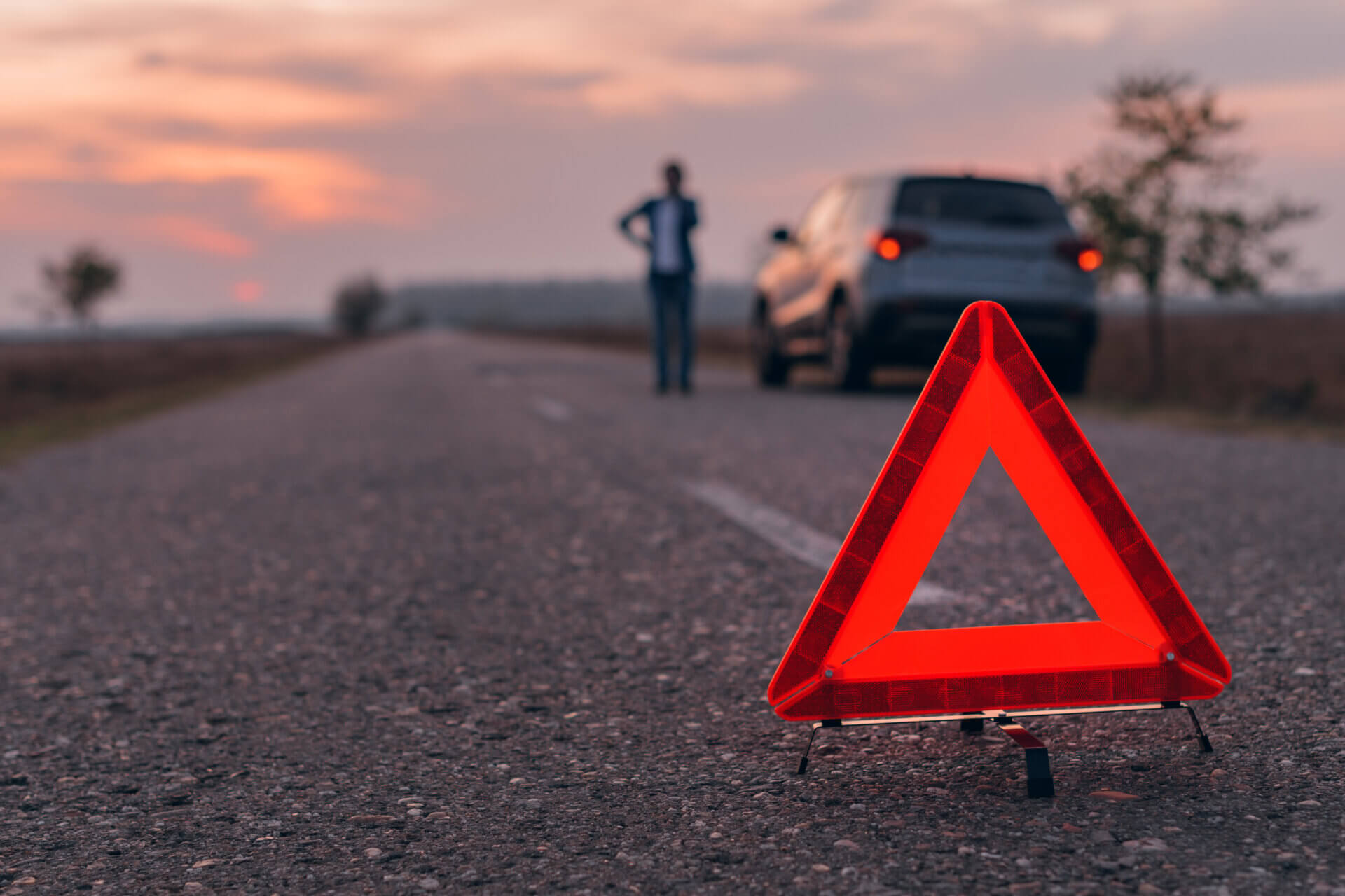 Destaque para um triangulo de sinalização de trânsito na rua. Ao fundo, uma pessoa está ao lado de um carro parado na estrada enquanto fala ao telefone.