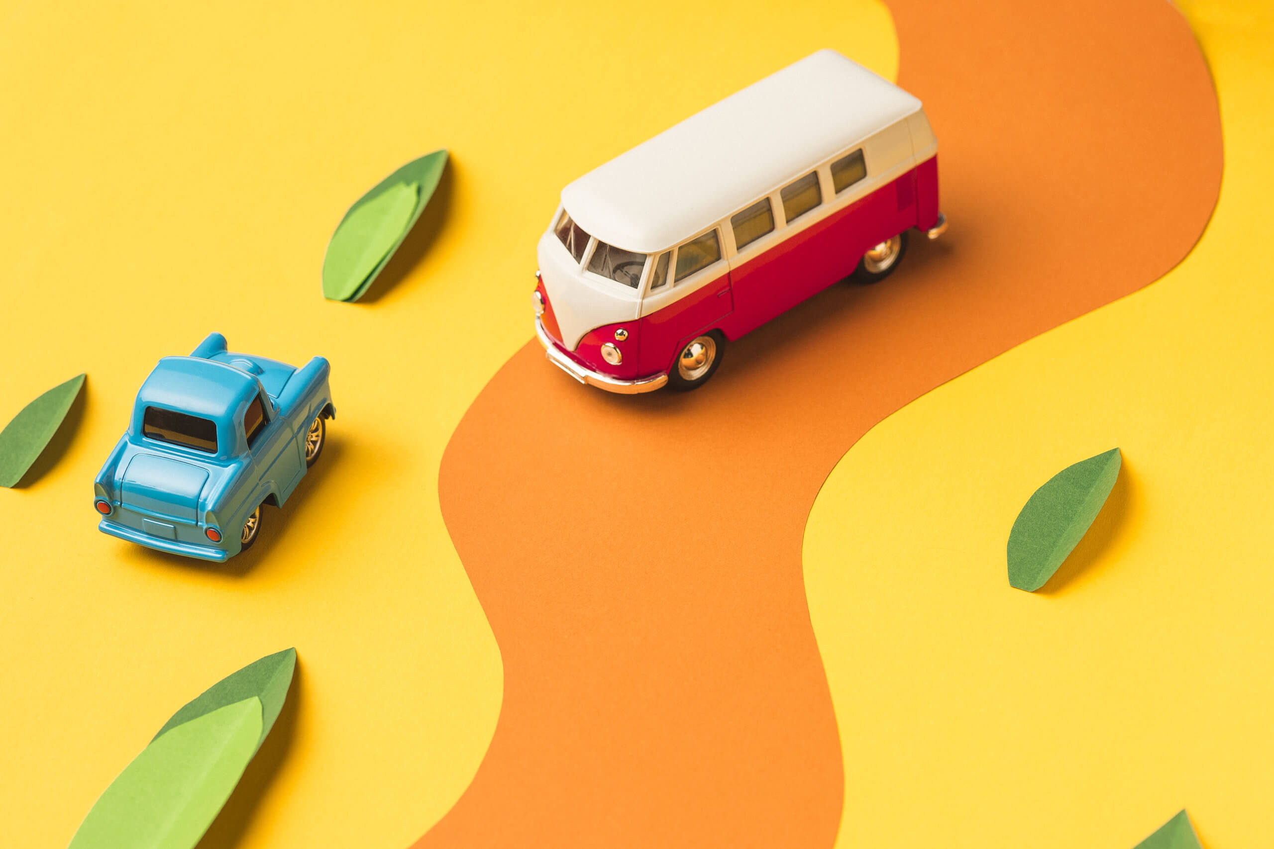 Ilustração de dois carros em miniatura, um azul e outro vermelho, em uma estrada sinuosa com folhas ao redor. 