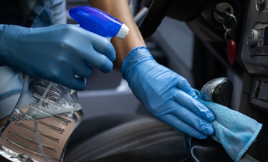 Mão com luva azul utilizando spray e pano para higienização de carro, focada na limpeza da alavanca de câmbio.