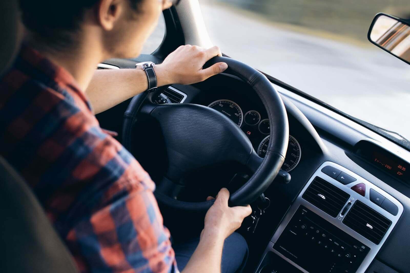 um homem está dirigindo um carro automático na estrada. Ele veste camisa xadrez vermelha e azul e tem um relógio preto no pulso esquerdo