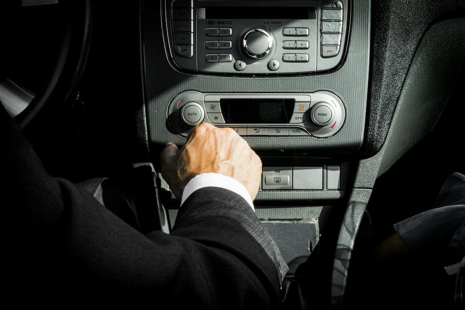 Mão de uma pessoa segurando a alavanca de câmbio de um carro, com o painel de controle de áudio e ar-condicionado ao fundo. A pessoa veste um terno preto.