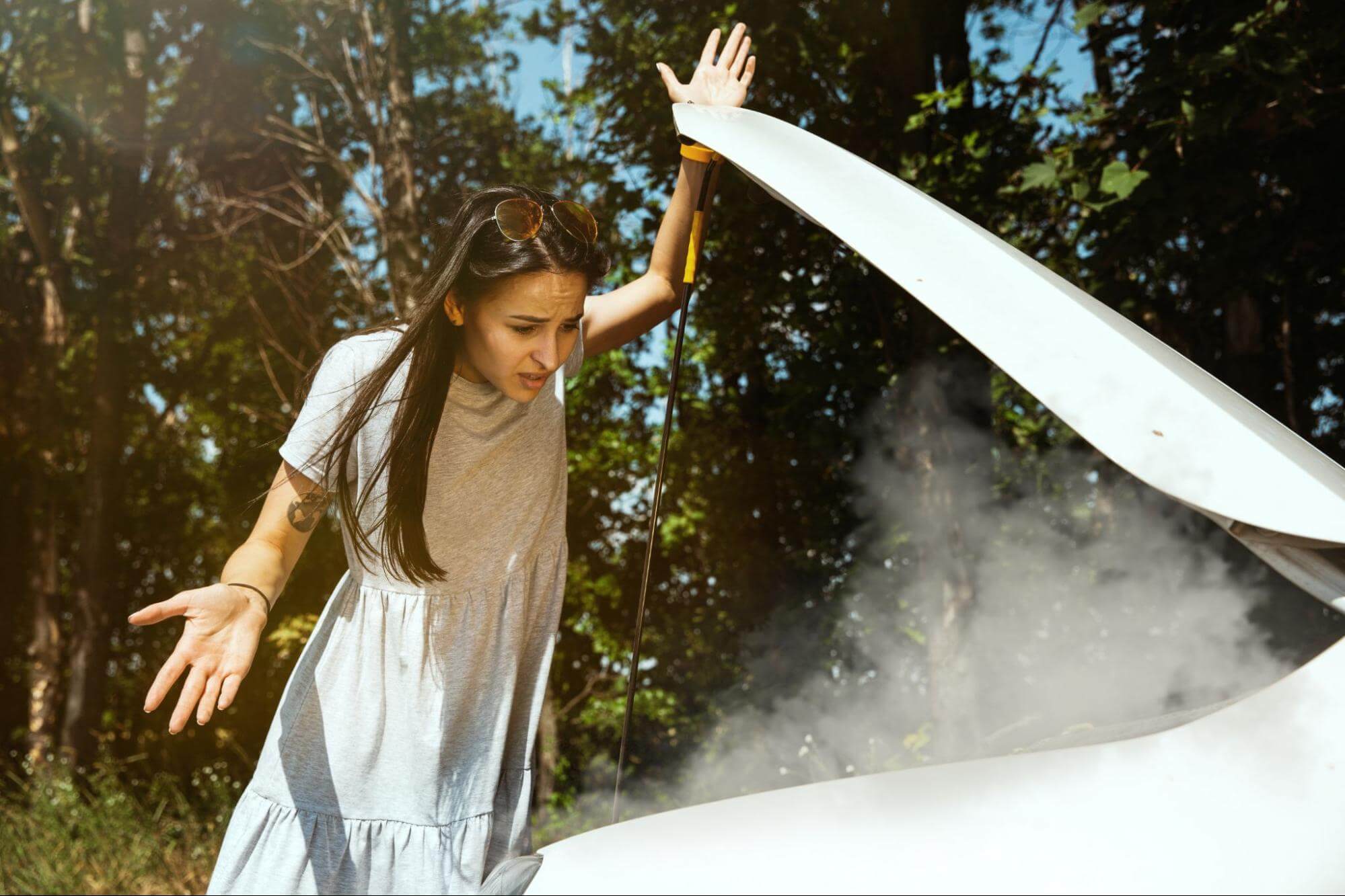 Na imagem, uma mulher branca, de cabelo castanho e liso está em frente a um veículo que está com o capô aberto, enquanto sai fumaça 