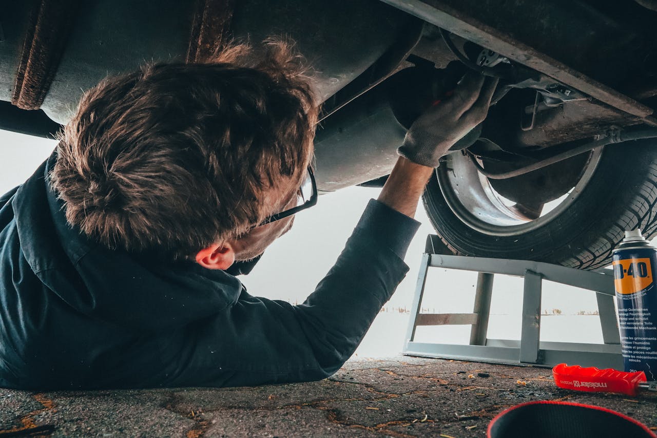 Mecânico deitado sob um carro, realizando uma inspeção detalhada na parte inferior do veículo, com ferramentas e produtos de manutenção ao lado.