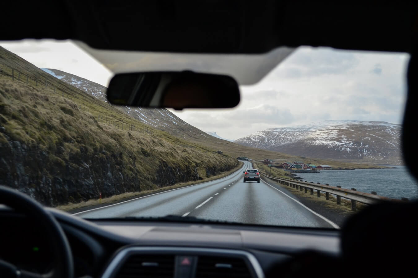 Vista de uma estrada sinuosa cercada por colinas e montanhas nevadas, vista de dentro de um carro em movimento.