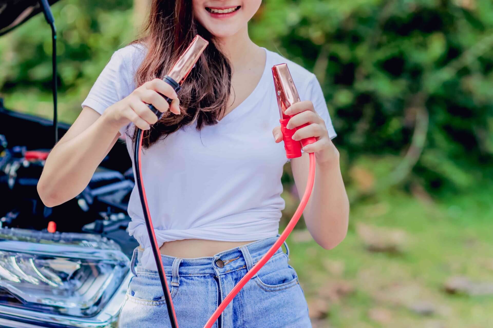 Mulher segurando dois cabos nas mãos, um positivo e um negativo, em frente a um carro com capô aberto.