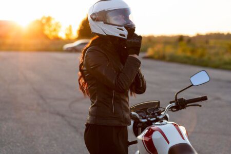 Motociclista de jaqueta de couro preta ajustando o capacete branco ao lado de uma moto, com o sol se pondo ao fundo.