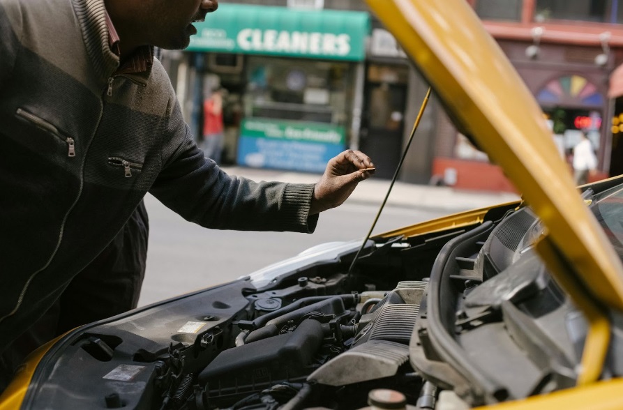 Homem inspecionando o motor de um carro amarelo, potencialmente verificando o corpo de borboleta e outros componentes essenciais para manutenção ou reparo, em um ambiente urbano.