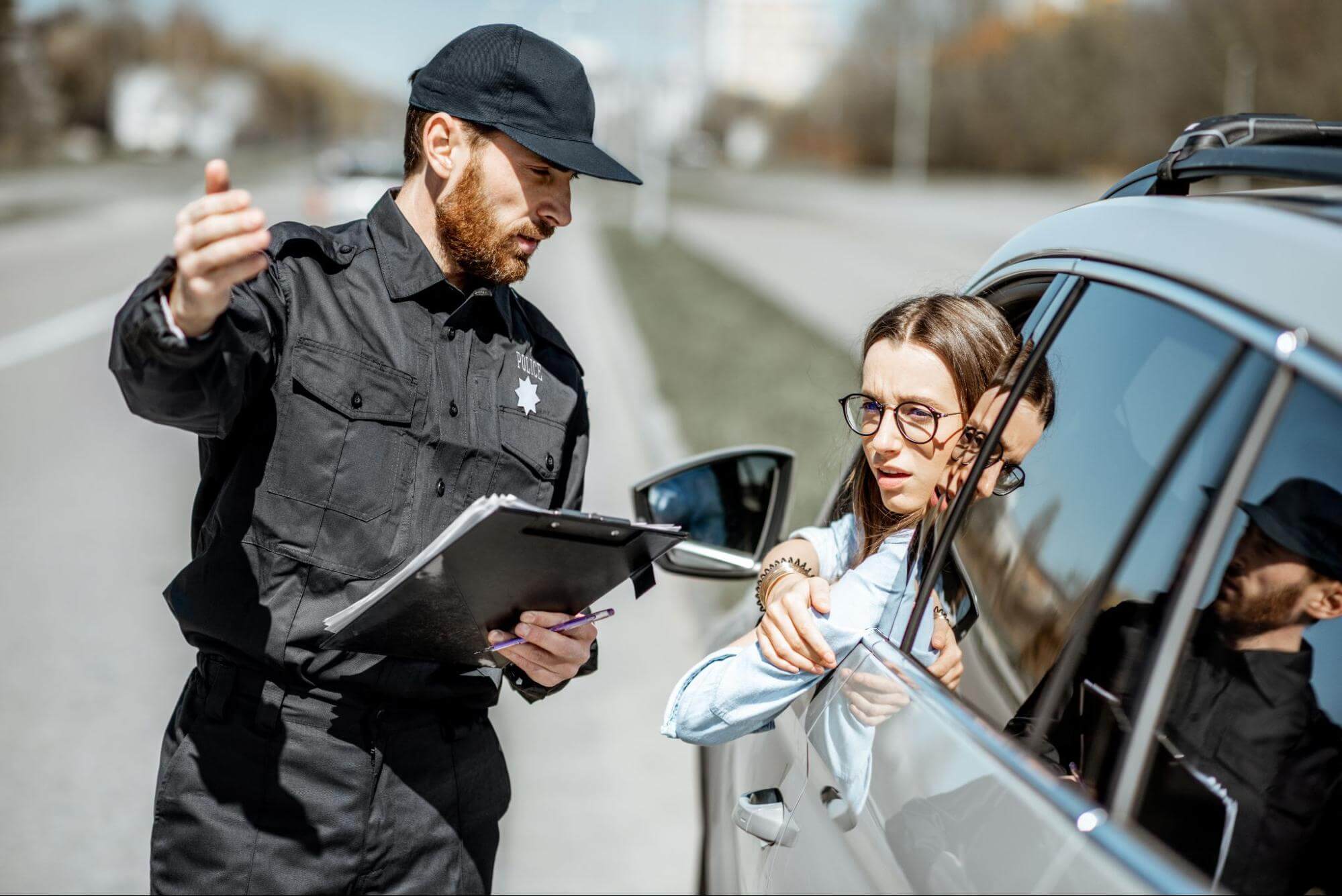 uma mulher está no lugar de motorista de um veículo, com a cabeça do lado de fora, enquanto conversa com um guarda de trânsito que tem uma caderneta em suas mãos e faz gestos com a mão, indicando direções