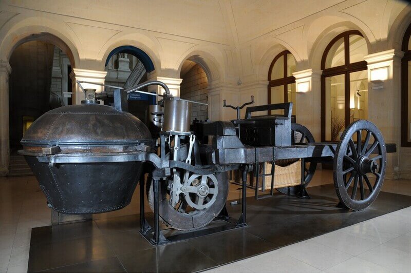 na imagem, encontramos um modelo do primeiro veículo a vapor, exposto como em um museu, em uma sala com espaço amplo e chão de madeira