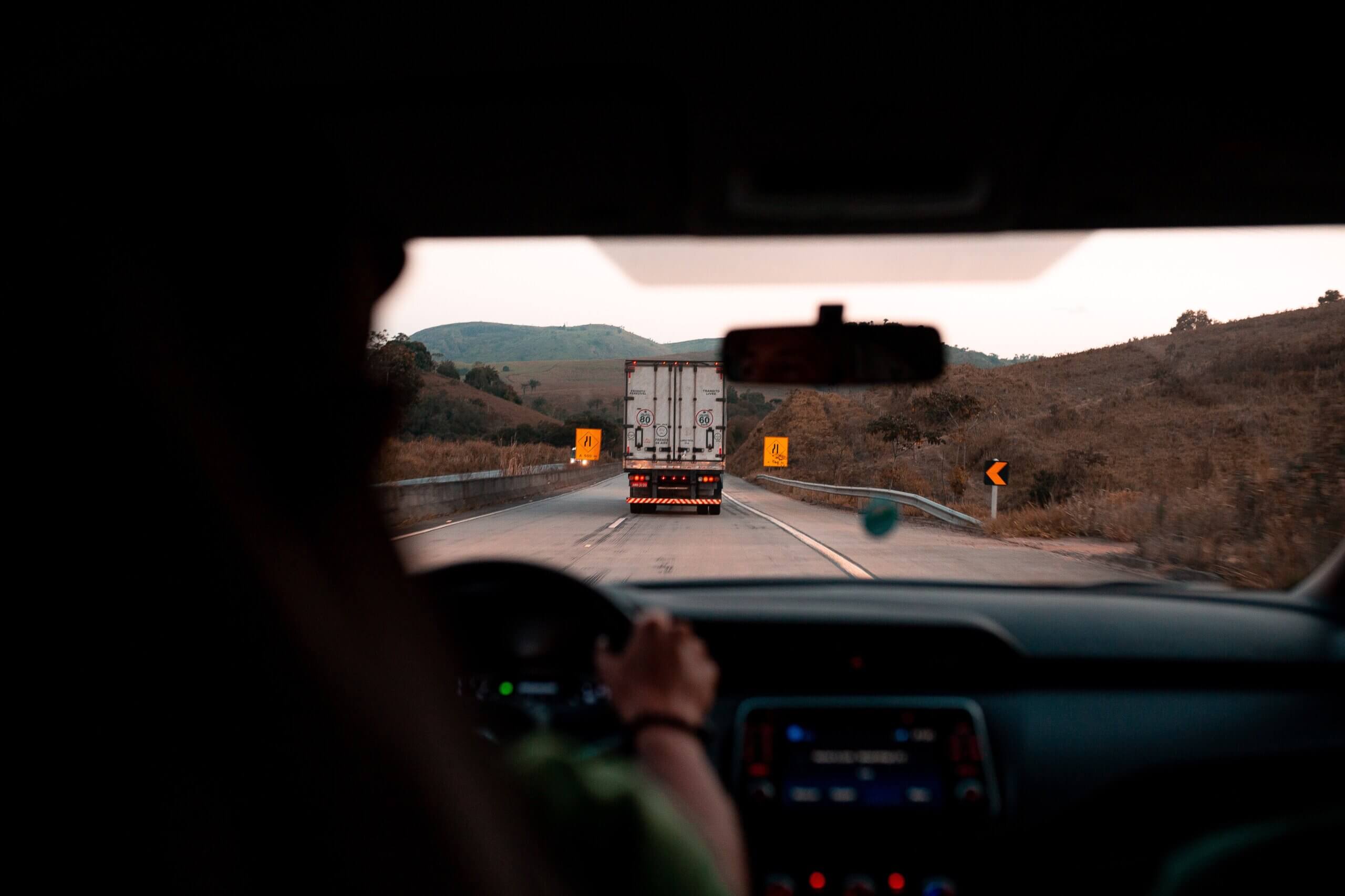 na imagem, vemos uma pessoa dirigindo um carro em uma rodovia, aparentemente ao final da tarde, e em sua frente está um caminhão e algumas placas de sinalização amarelas