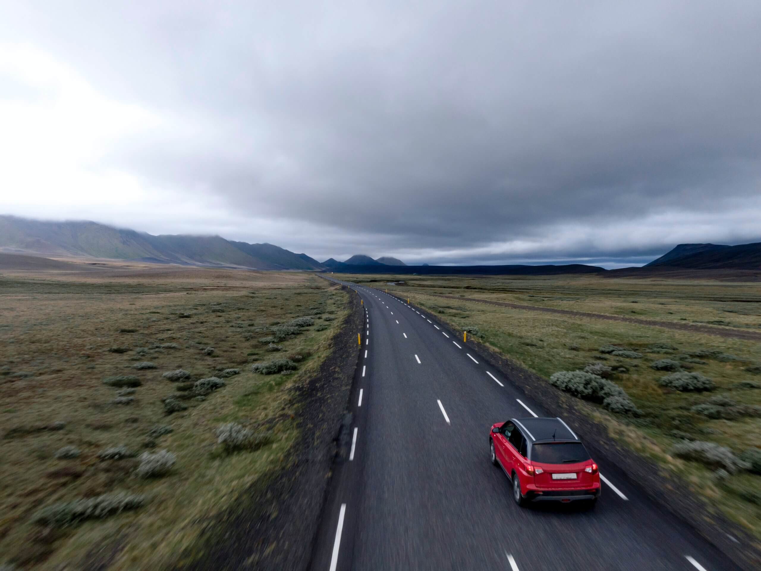 Um carro vermelho está em movimento em uma pista de duas faixas, na qual ele é o único veículo à vista. Ao redor da pista é possível ver vegetações baixas. No horizonte estão algumas nuvens e um terreno montanhoso 
