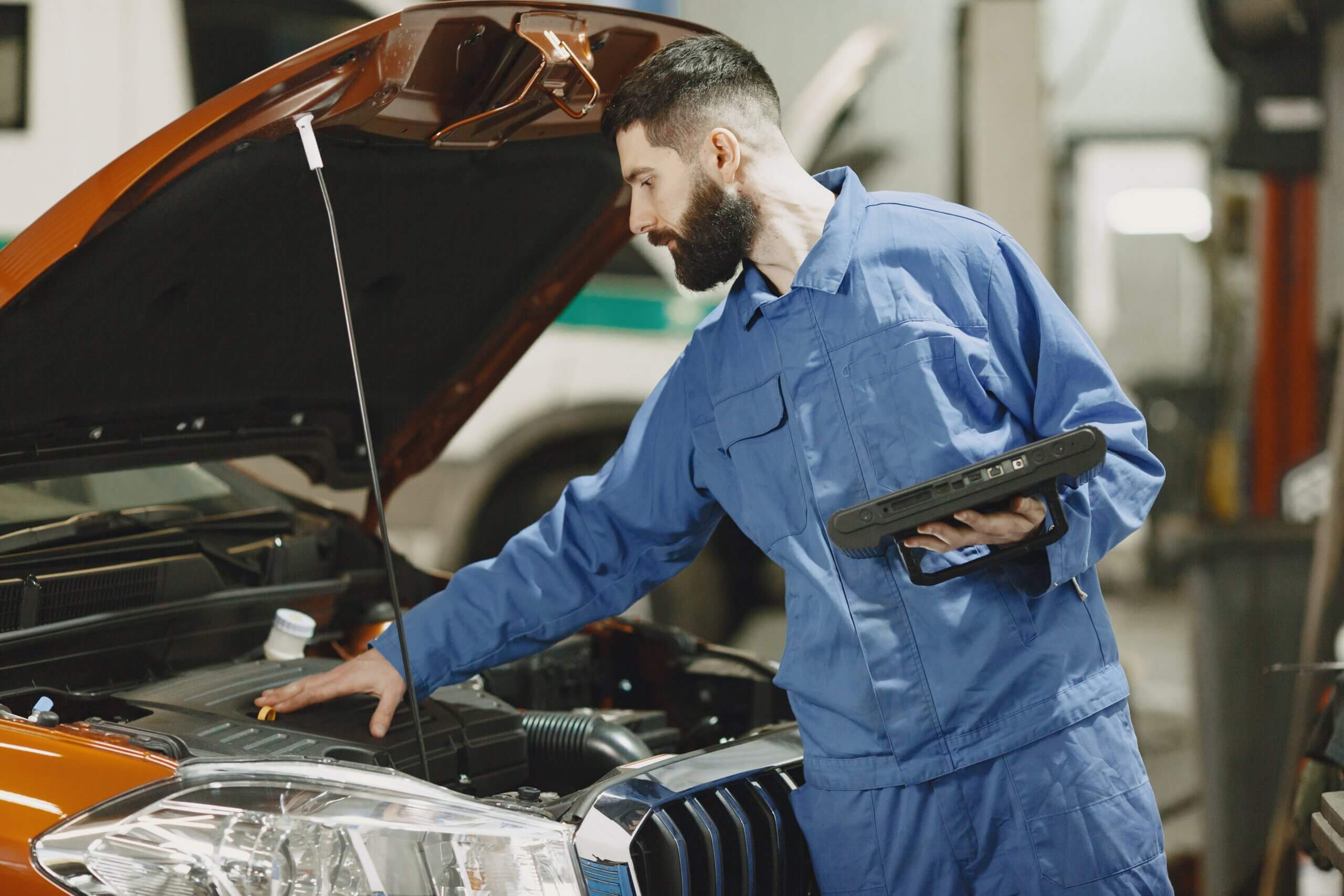um homem que veste roupas de mecânico está segurando uma peça automotiva enquanto, com a outra mão, mexe no interior do capô de um carro.