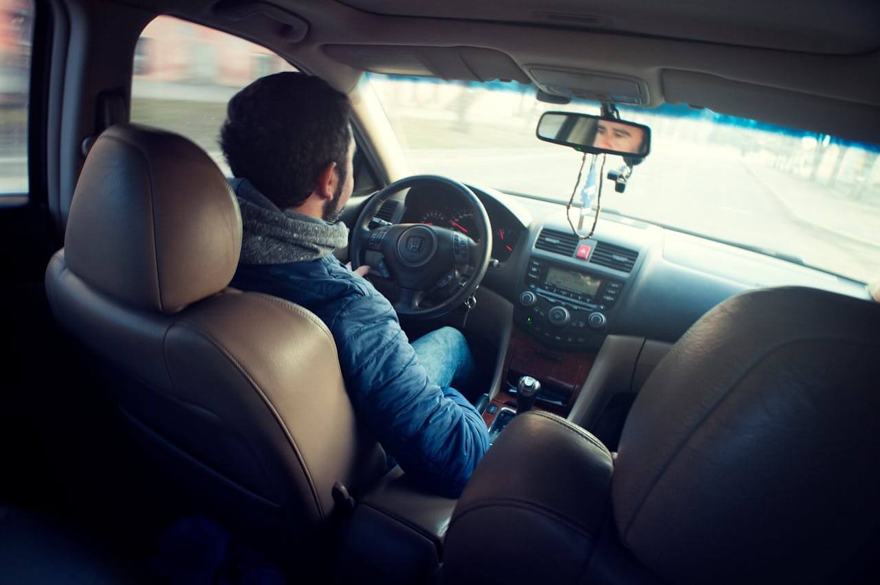 Homem dirigindo um carro em uma estrada, visto de trás. O interior do carro é moderno e bem conservado.