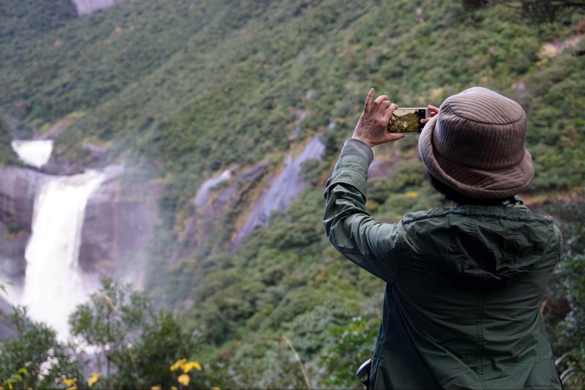 uma mulher que veste blusa de manga comprida verde e um chapéu está de costas para a imagem enquanto tira fotos de uma cachoeira presente em uma montanha que está à sua frente