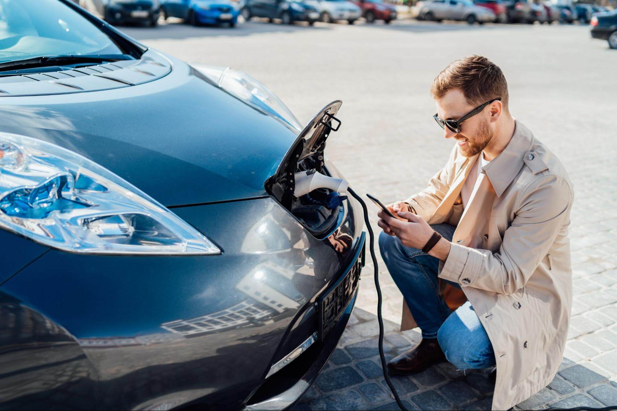 um homem com calça jeans e blazer está abaixado próximo ao seu carro de motor elétrico enquanto aguarda seu carregamento via rede elétrica. Ele mexe em seu celular e sorri.