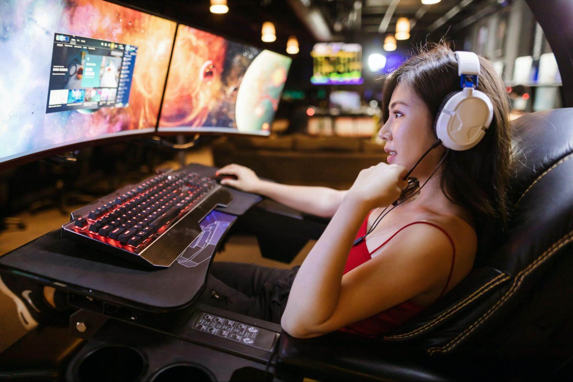 uma mulher que veste uma regata vermelha e usa fones de ouvido está sentada em uma cadeira gamer, à frente de um setup com diversos monitores e um teclado com luzes coloridas.