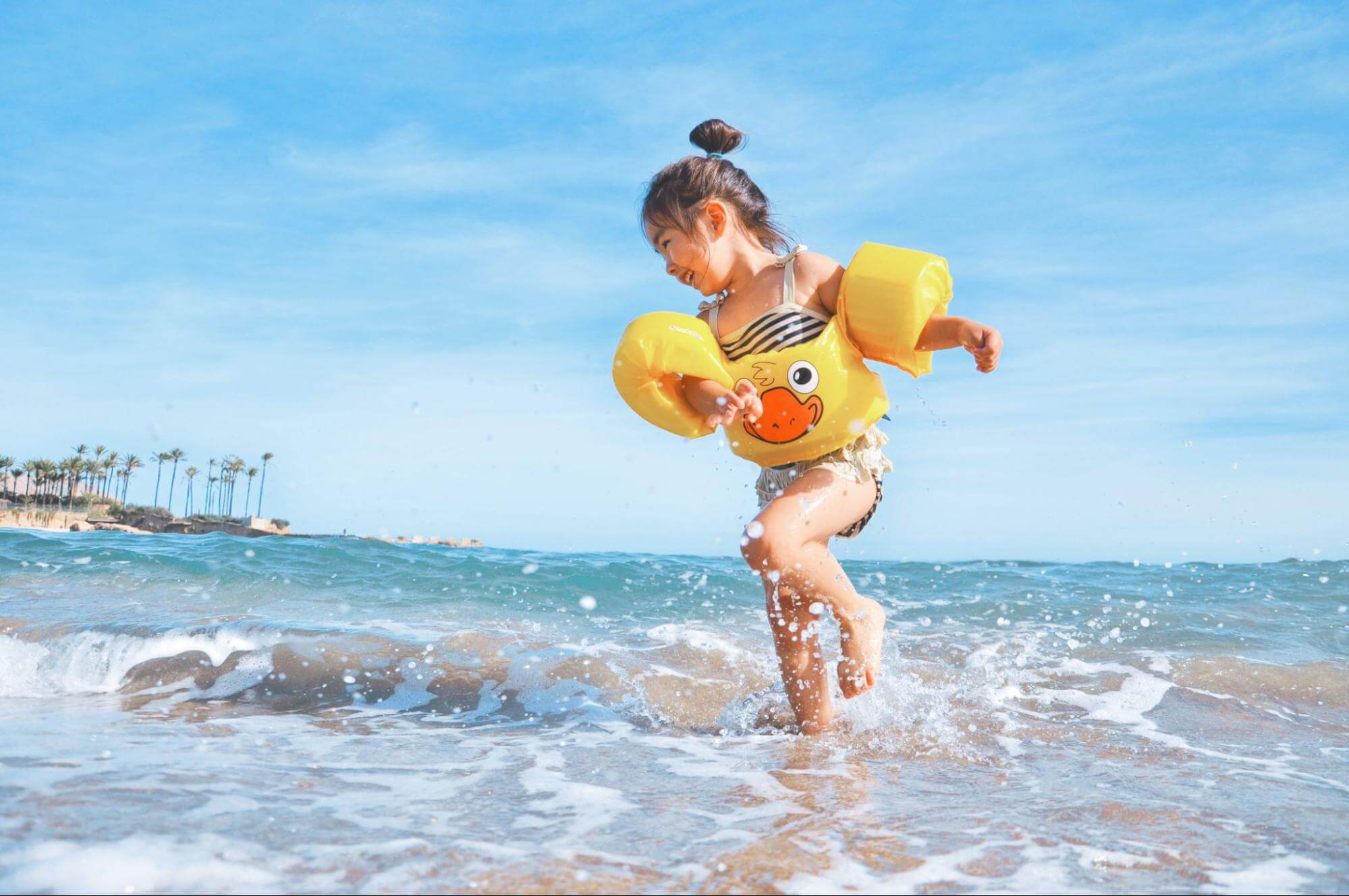 uma menina de aproximadamente 3 anos está brincando na praia, com os pés no mar. Ela veste um maiô colorido e boias nos braços, e está sorrindo enquanto pula ondas