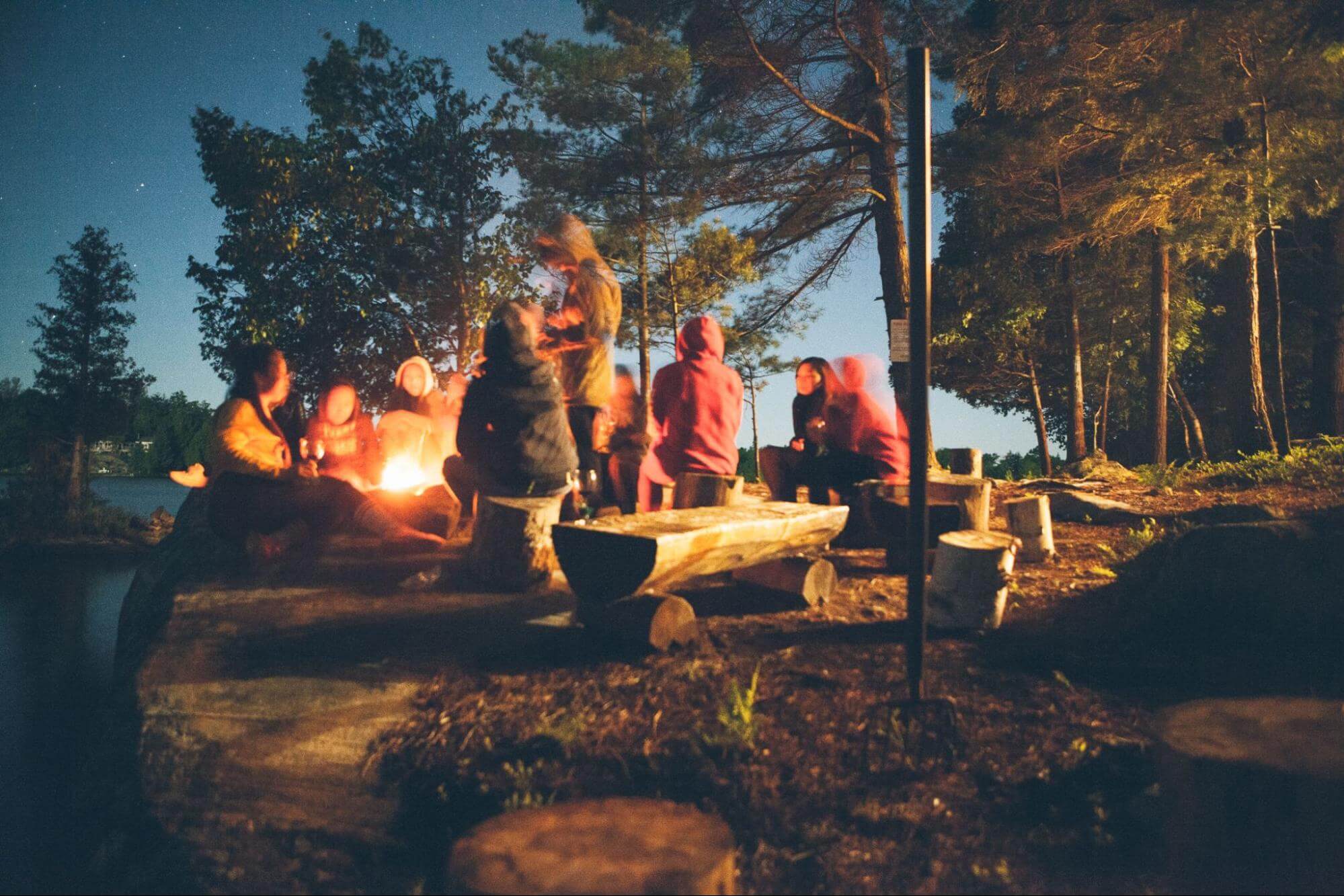 um grupo de pessoas está em um camping, reunidas ao redor de uma fogueira enquanto conversam.