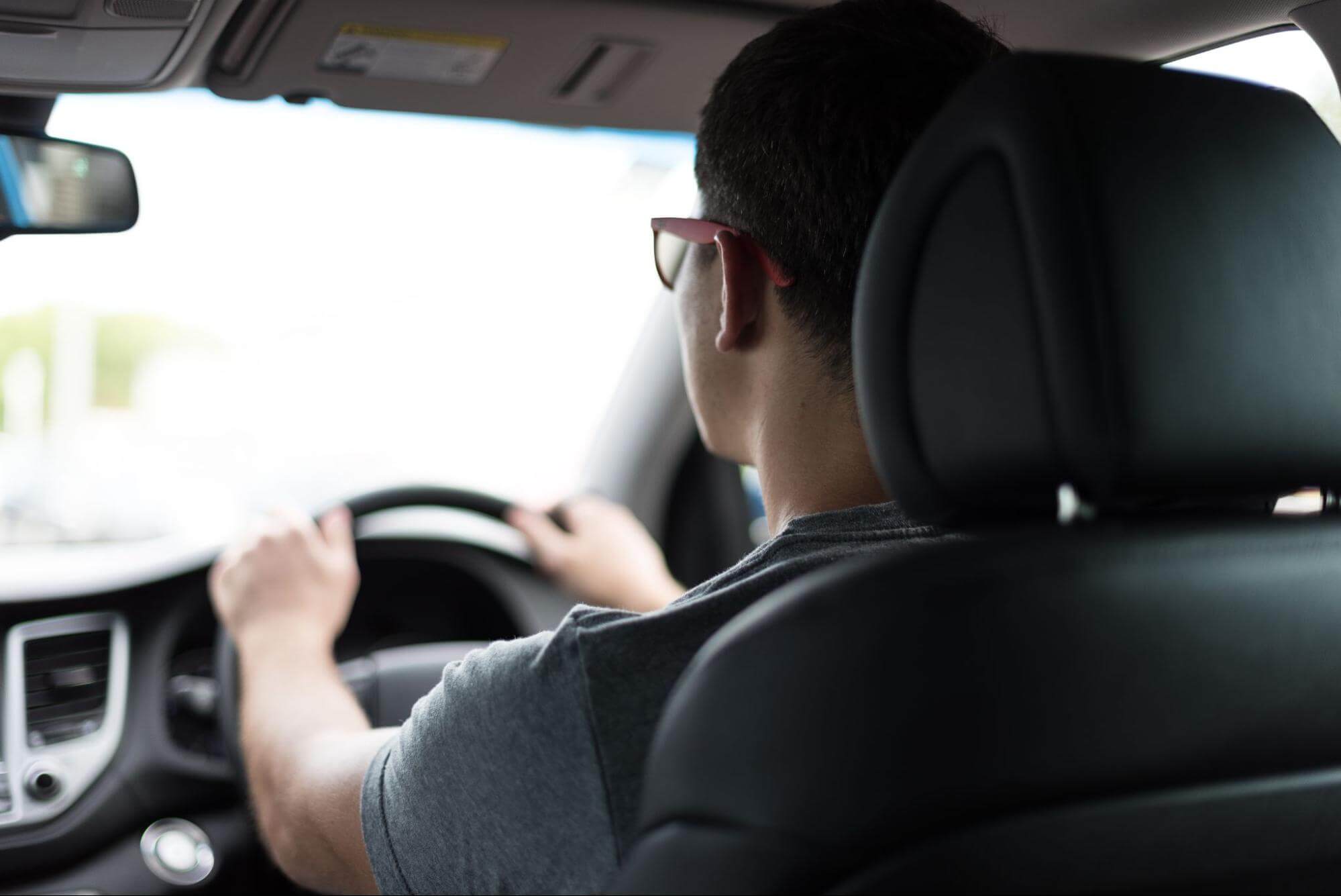 um homem está dirigindo um carro, com as mãos no volante. Ele usa uma camiseta cinza, óculos de grau e está com o cinto de segurança acionado