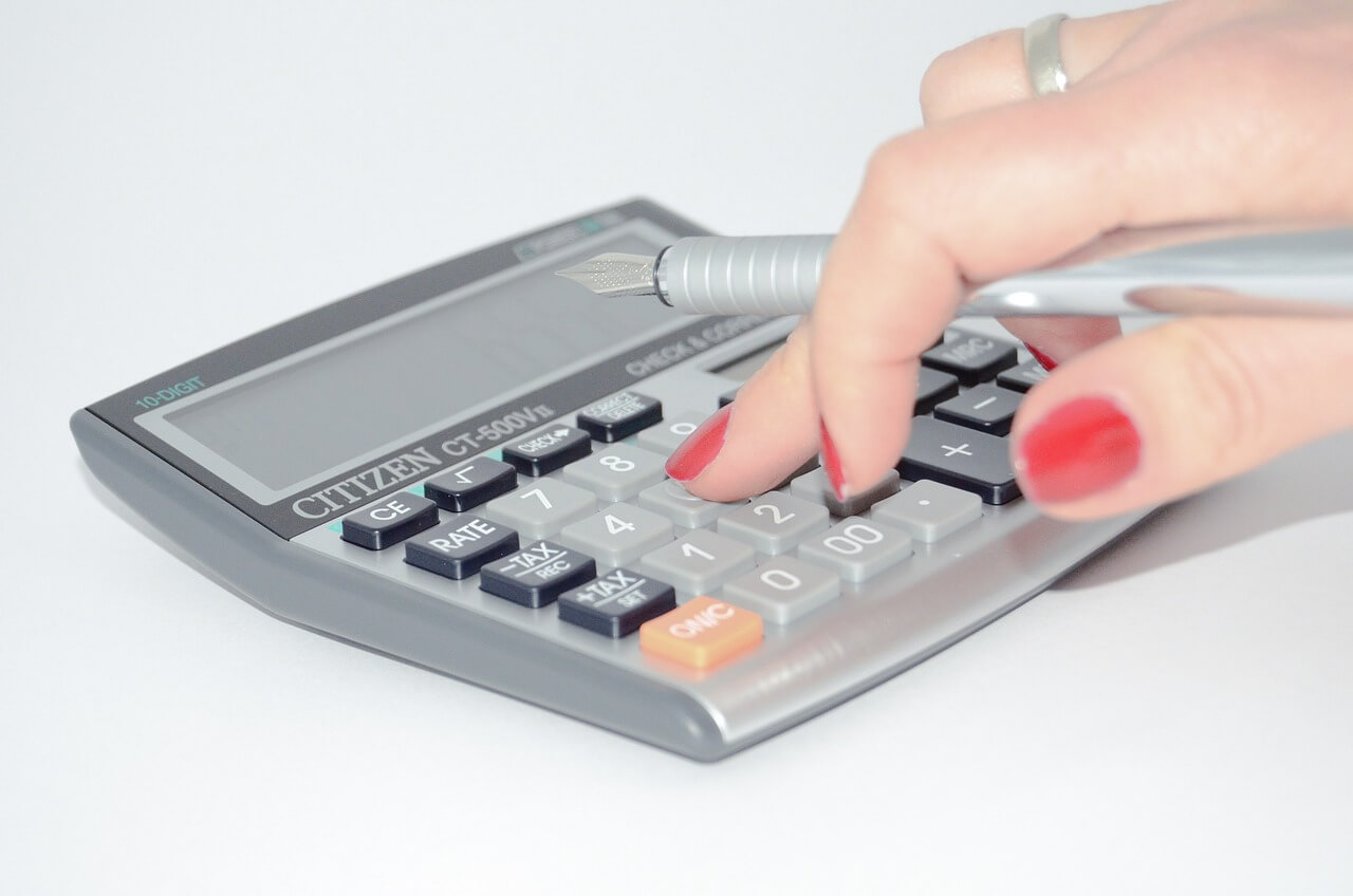 uma mão feminina está digitando em uma calculadora analógica. Ela tem as unhas pintadas de vermelho, usa um anel em prata e segura uma caneta entre os dedos