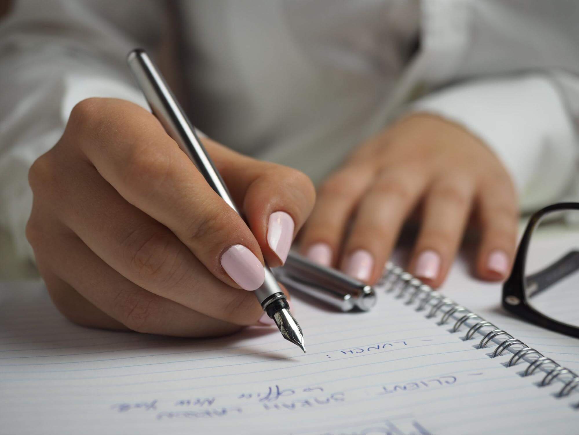 uma mão feminina está escrevendo em um caderno, fazendo contas. Ela tem as unhas pintadas de pérola e, em desfoque ao fundo, é possível ver que ela veste trajes de escritório cinza