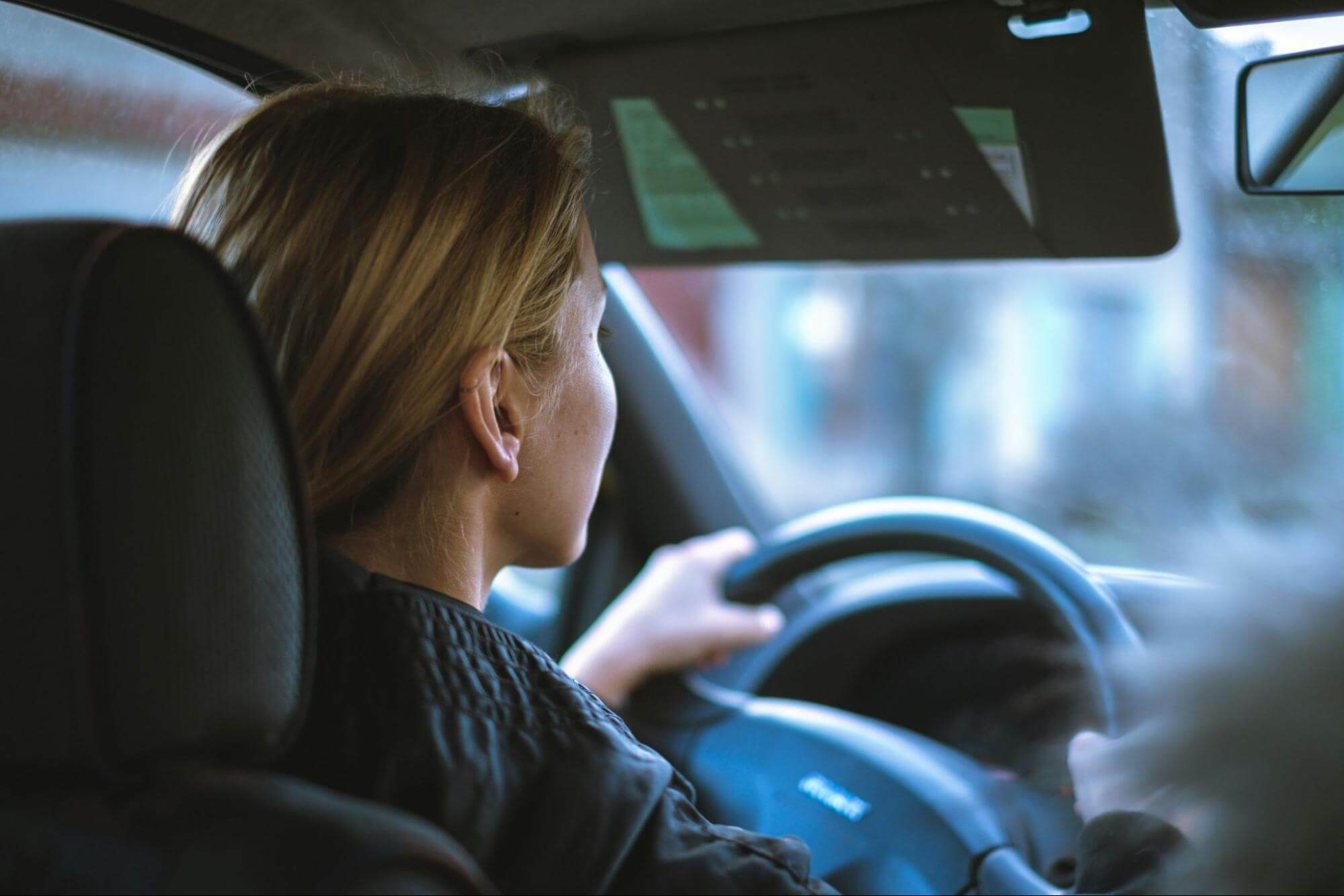 uma mulher loira, de cabelos presos, está dirigindo um carro na rua tranquilamente