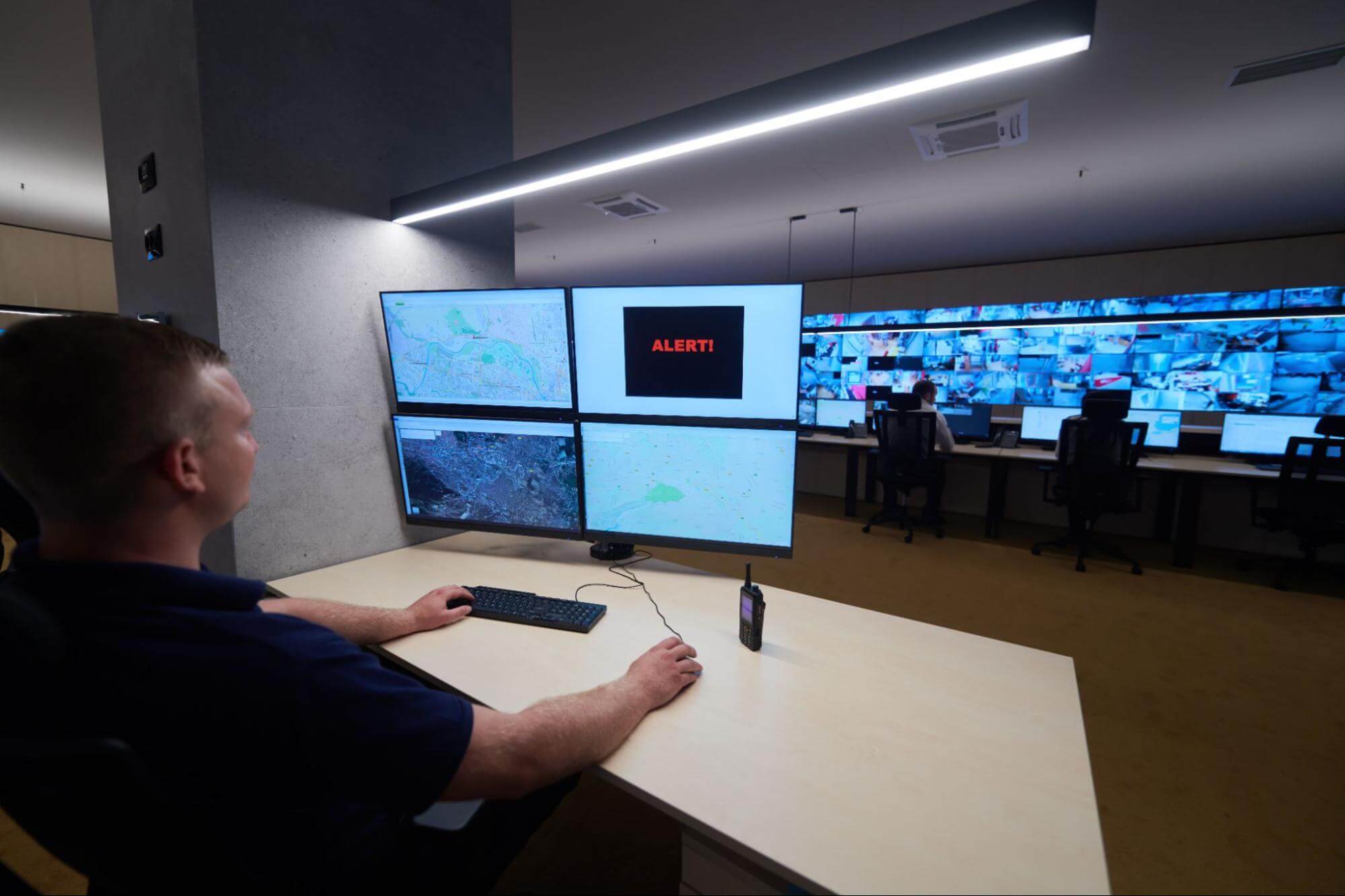 um profissional de segurança está em uma sala com diversos monitores, onde é possível notar o acompanhamento remoto de diversos ambientes diferentes