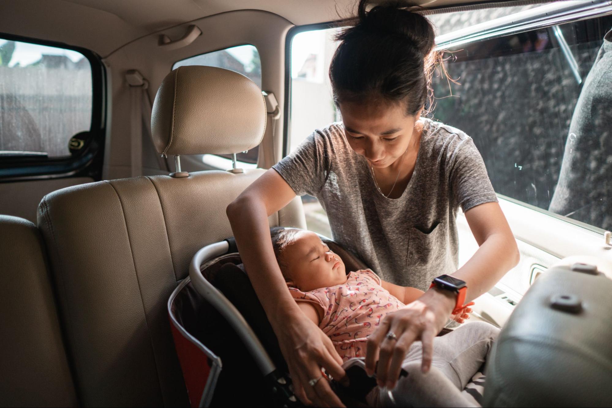 uma mulher está em pé ao lado do carro que tem sua porta aberta. No banco traseiro está uma cadeirinha bebe conforto com um bebê. A mulher está acertando as configurações da cadeirinha para o bebê.