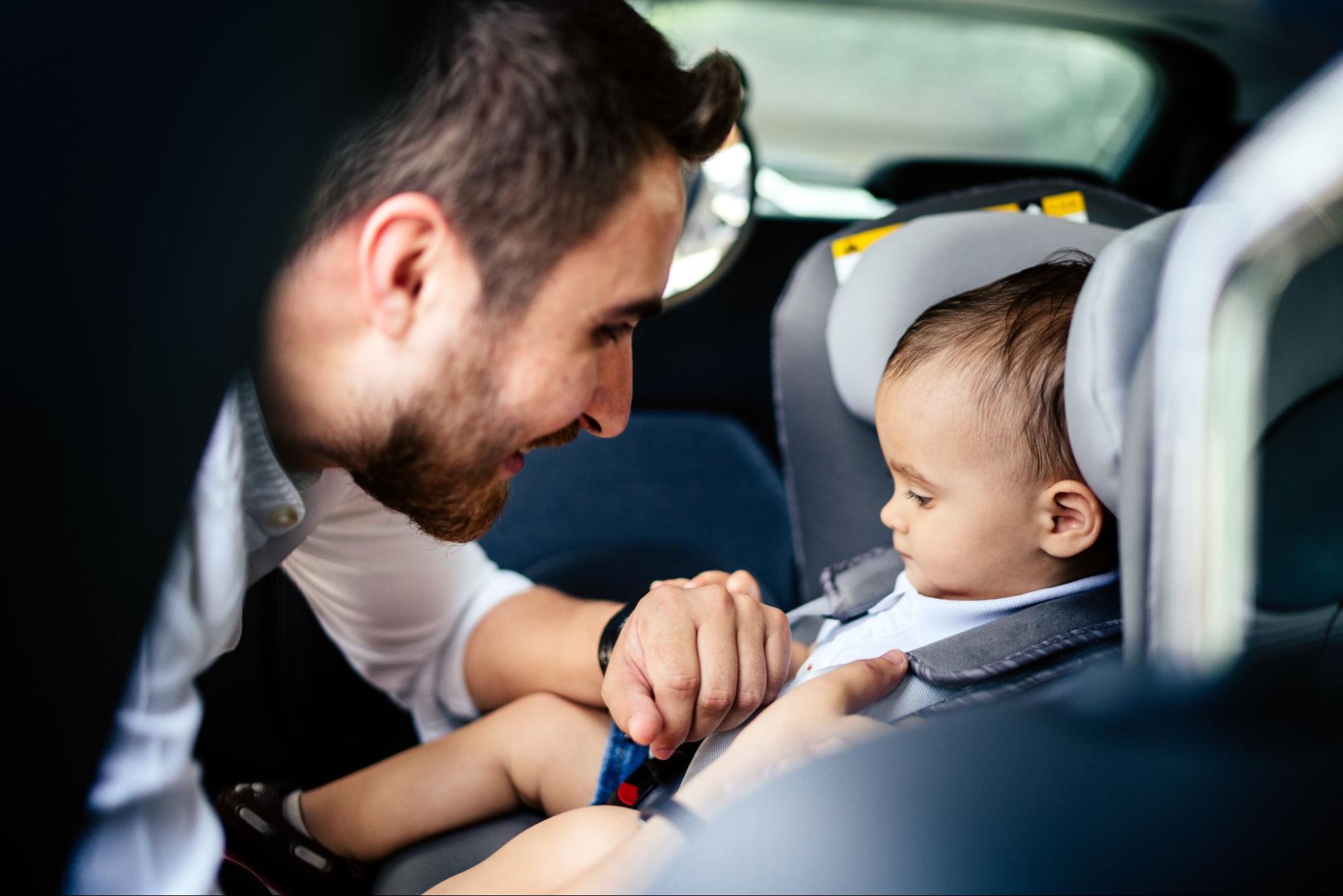 um homem com barba está configurando uma cadeirinha no banco de trás de um veículo. Nela, está sentado um bebê.