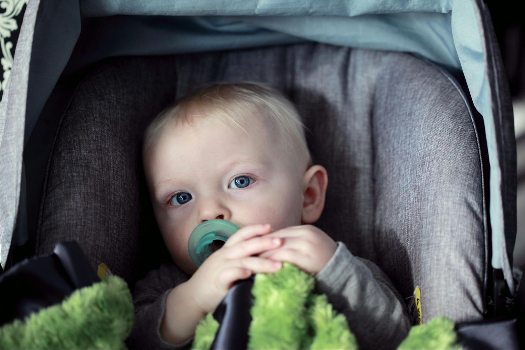 um bebê com cabelos loiros e roupa verde está sentado em uma cadeirinha. Ele tem uma chupeta na boca e segura um dos cintos da cadeirinha com as duas mãos
