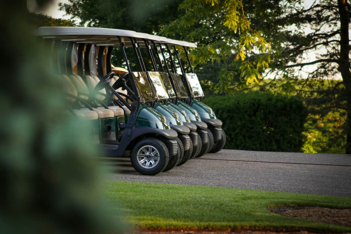 Uma fileira de carrinhos de golfe estacionados ao longo de uma trilha pavimentada em um campo de golfe, com árvores e arbustos ao redor. Os carrinhos são de cor verde escura e equipados com telhados brancos. Estes veículos são alimentados por baterias de tração monobloco, garantindo eficiência e confiabilidade.