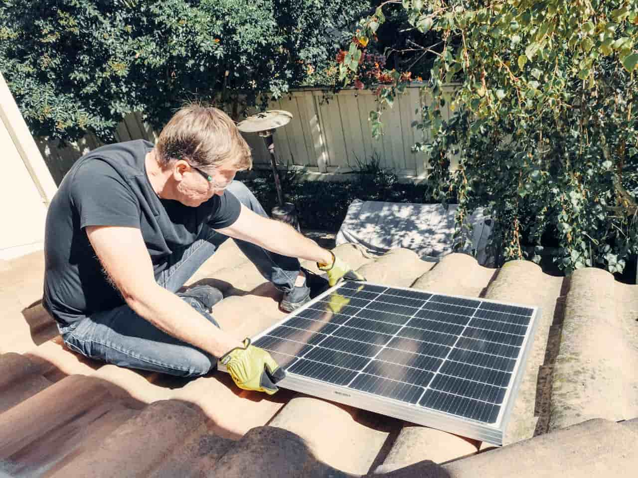 Pessoa instalando um painel solar em um telhado, usando luvas de proteção. A imagem mostra um painel solar sendo posicionado no telhado, com árvores e vegetação ao fundo.