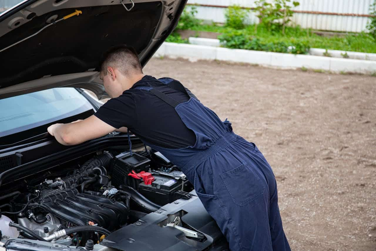 Mecânico de macacão azul inspeciona o motor de um carro com o capô aberto, concentrando-se na bateria e nos componentes ao redor em um ambiente externo.