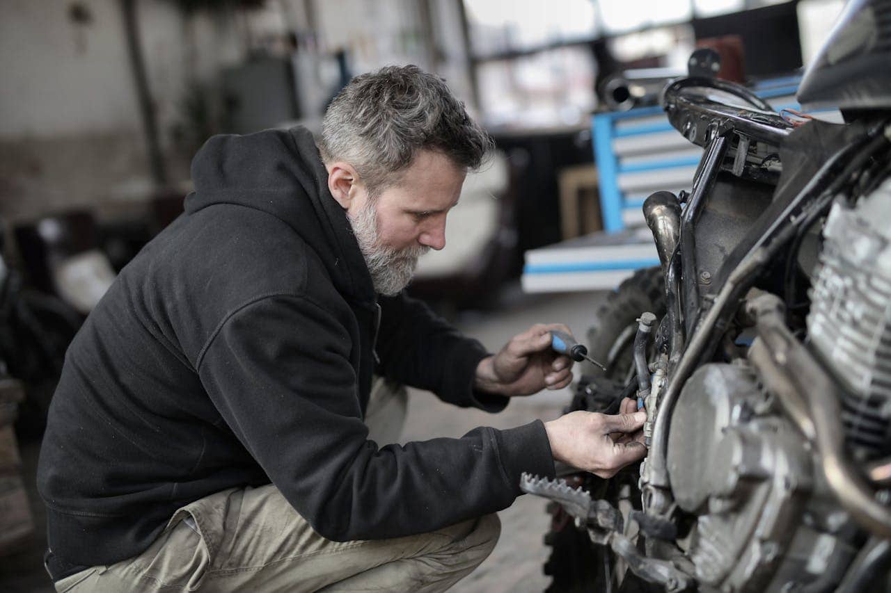  Mecânico de barba grisalha, usando moletom preto, trabalha na manutenção do motor de uma moto em uma oficina, utilizando uma chave de fenda no reparo da ignição da moto.