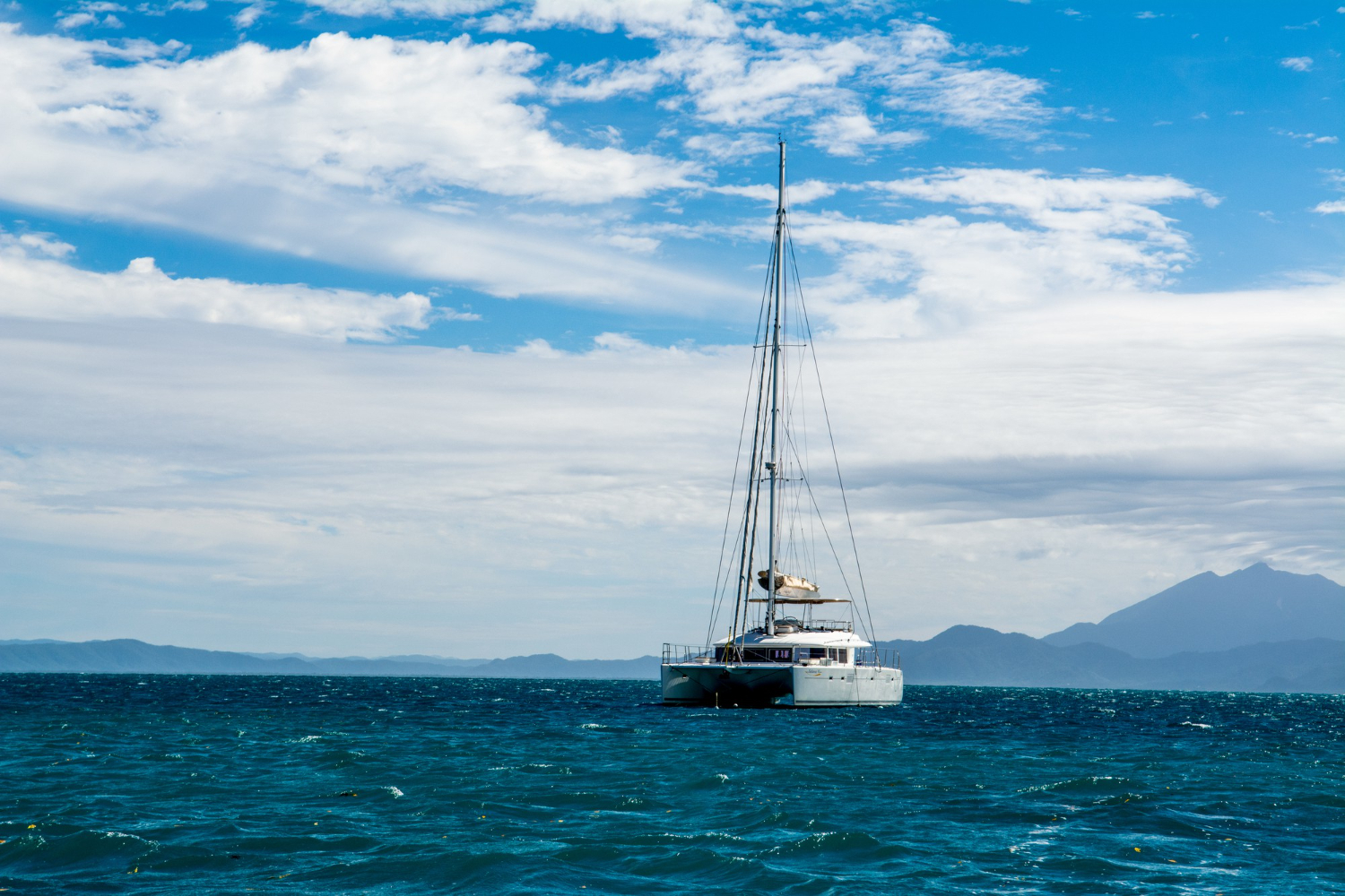 Imagem de um catamarã navegando em mar aberto, com um céu azul e nuvens brancas acima. Montanhas são visíveis no horizonte ao fundo.