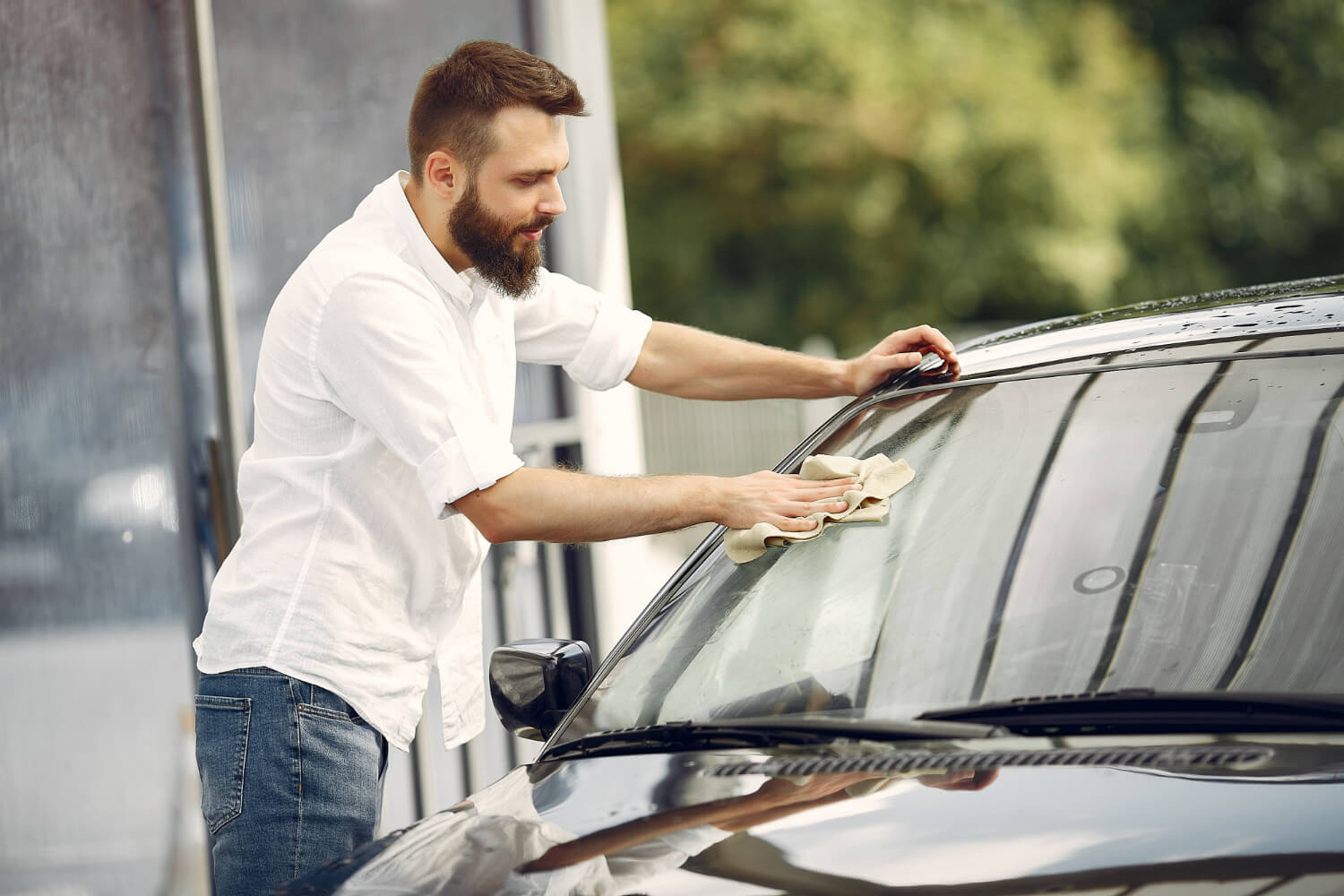 Homem limpando o para-brisa de um carro preto com um pano, ao ar livre, em um dia ensolarado. Ele está usando uma camisa branca e jeans, concentrado na tarefa.