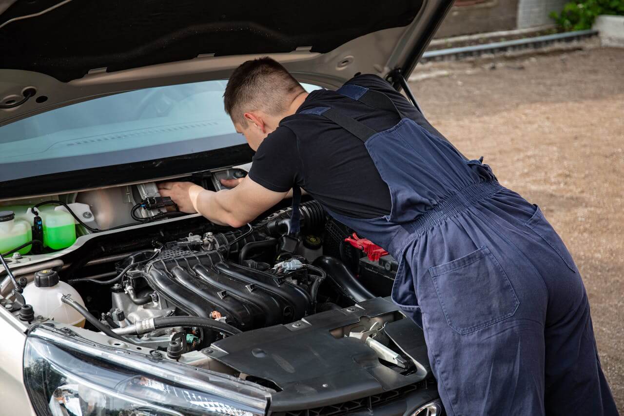 Mecânico ajustando o motor de um carro com o capô aberto, mostrando componentes do sistema elétrico e da bateria.