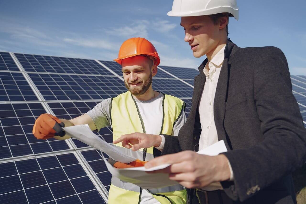 ois engenheiros, um com capacete laranja e colete de segurança, e outro com capacete branco e terno, analisam documentos e discutem em frente a painéis solares em um campo de energia solar.