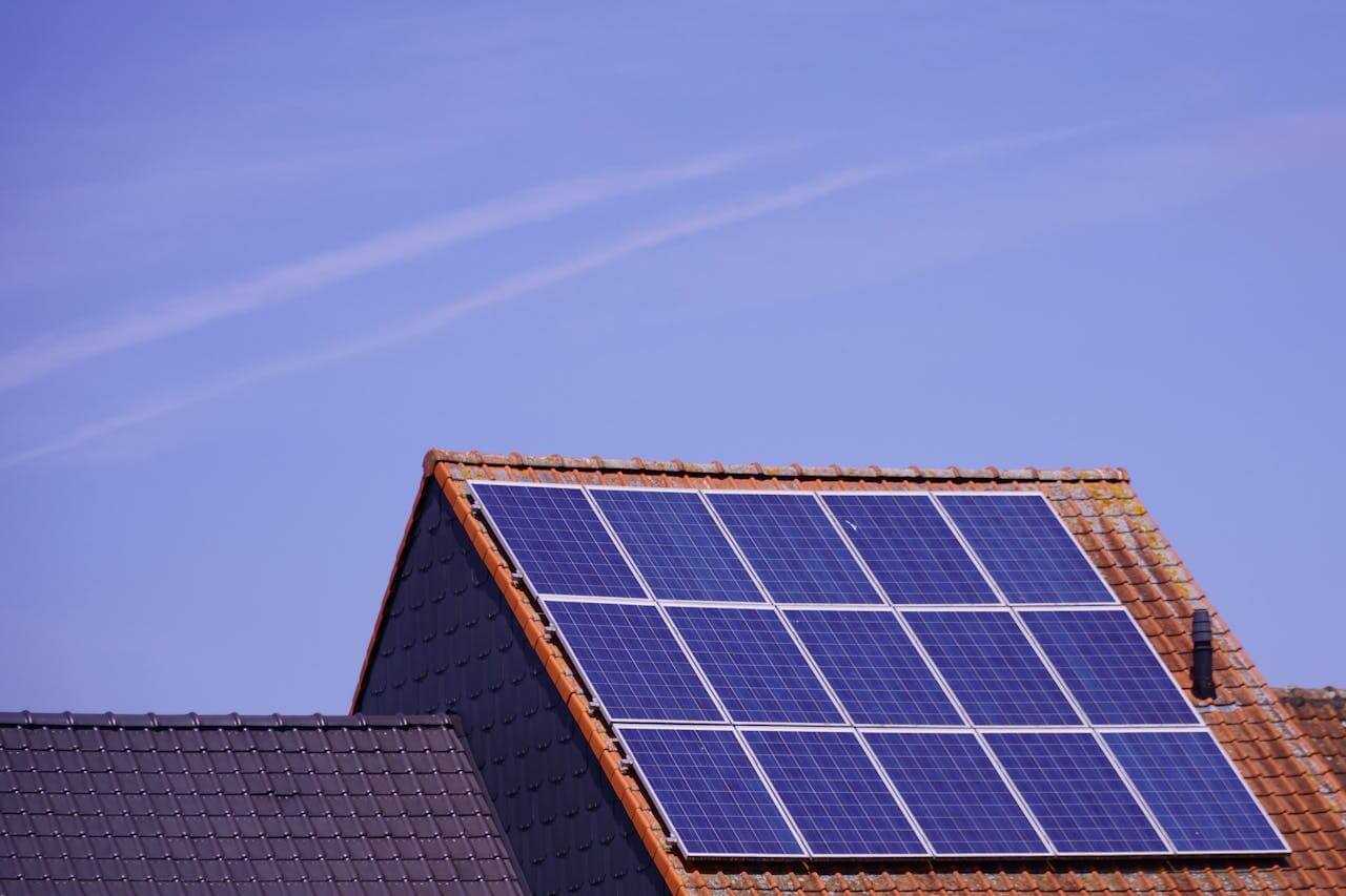 Telhado de uma casa com telhas de cerâmica vermelhas, equipado com paineis solares que captam energia solar sob um céu azul claro.