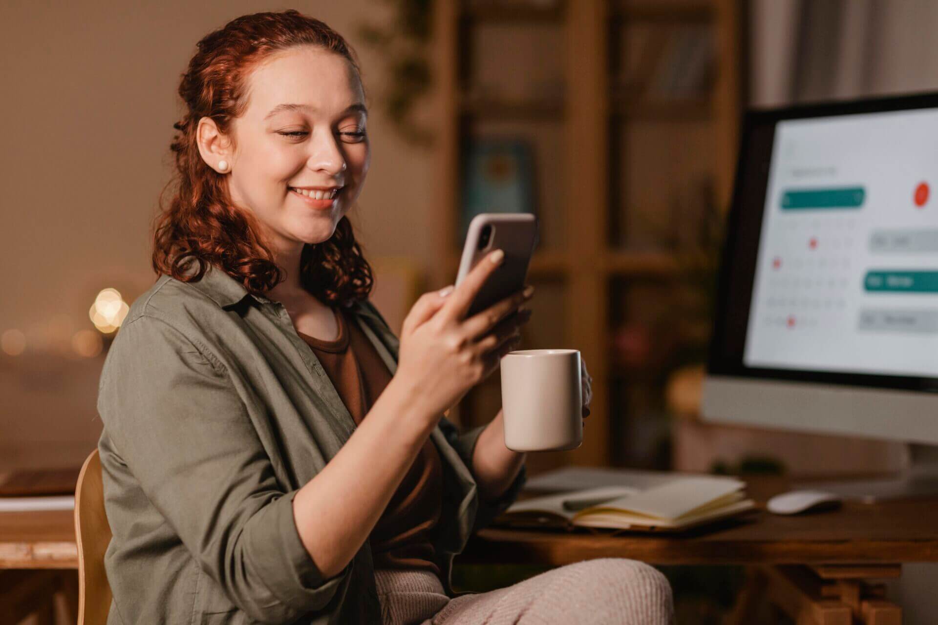 Mulher sorridente segurando um smartphone com uma mão e uma xícara de café com outra, enquanto está sentada em frente à uma mesa com um desktop.