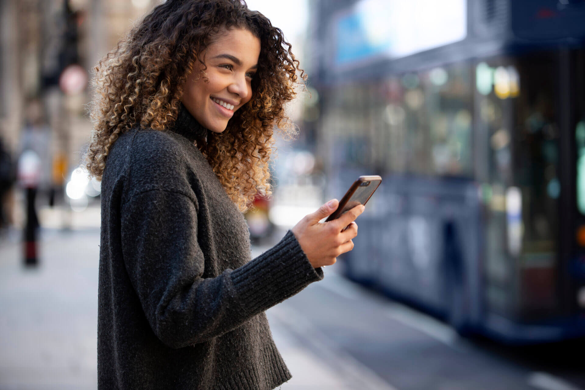 Mulher com um smartphone na mão enquanto está parada na calçada de uma avenida movimentada.