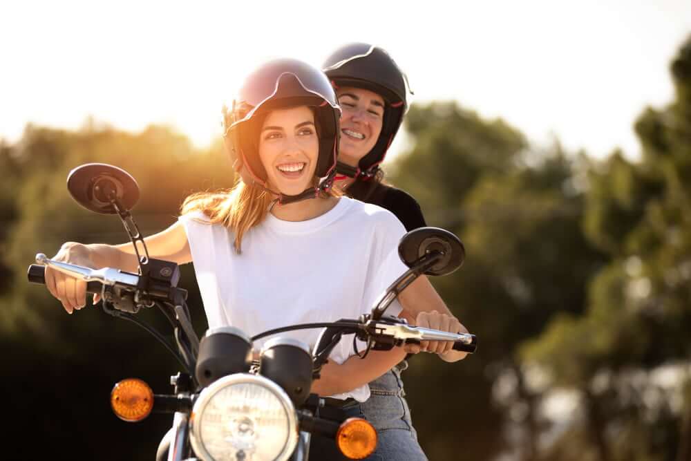 Duas pessoas sorrindo em uma motocicleta, usando capacetes, em um ambiente ao ar livre com luz do sol.