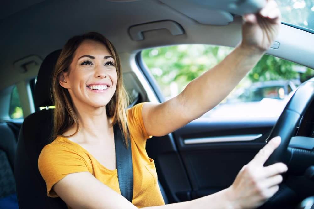 Mulher sorridente dirigindo um carro, com uma mão no volante e a outra ajustando o espelho.