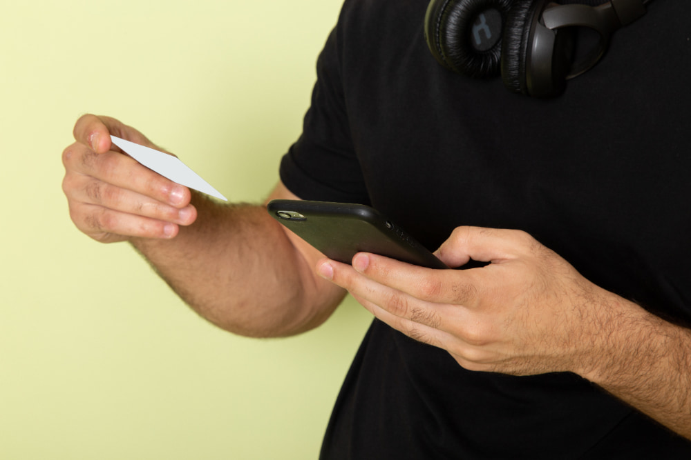 Homem segurando um cartão branco enquanto utiliza um smartphone.