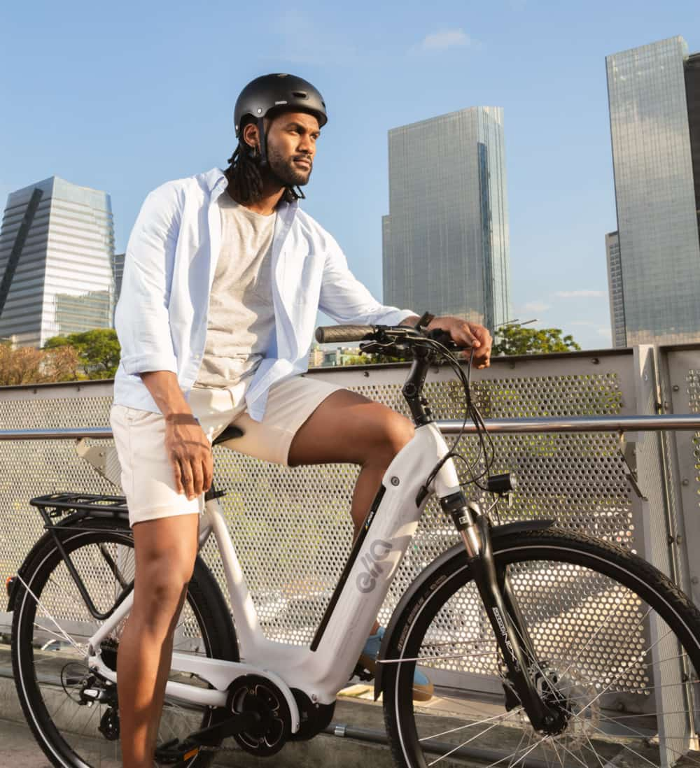 Homem com capacete, sentado em uma bicicleta elétrica Ella, com edifícios modernos ao fundo e céu azul.