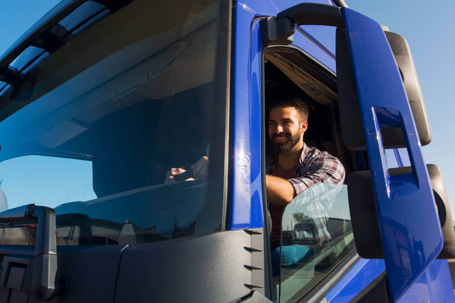 Um homem com barba está sentado na cabine de um caminhão azul, olhando para fora pela janela aberta. Ele está sorrindo e vestindo uma camisa xadrez. O céu está claro ao fundo, indicando um dia ensolarado.