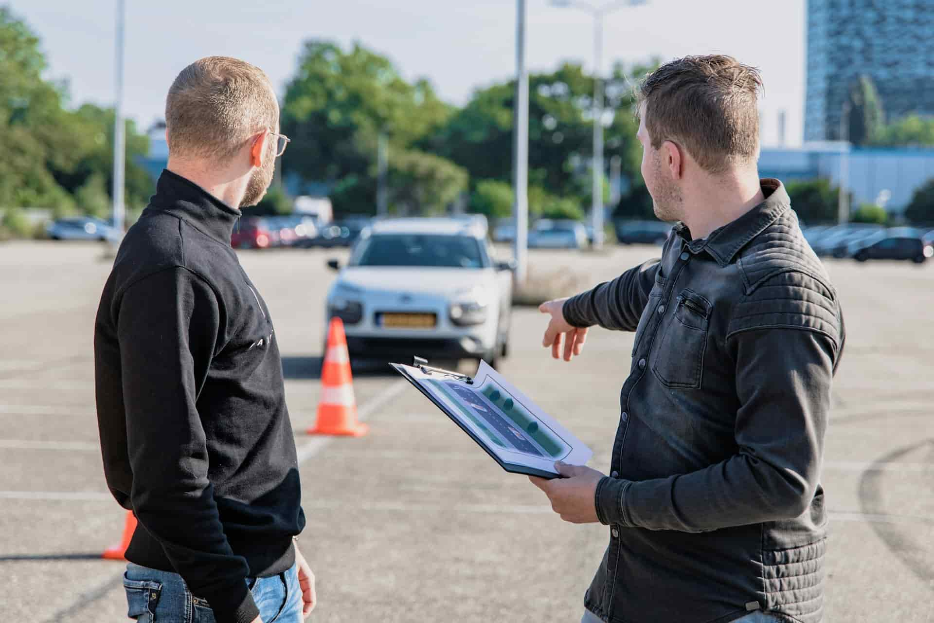 Dois homens conversam em um estacionamento. Um homem, de costas, usa um suéter preto e observa enquanto o outro, vestindo uma camisa cinza, aponta para um carro branco à frente e segura uma prancheta com documentos. Cone laranja está visível ao fundo.